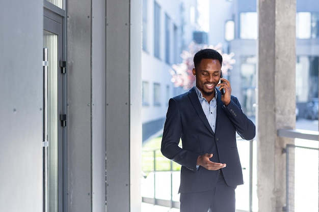 Successful employee smiles and talks on the phone, rejoices, African American male businessman, near the office outside, with a mobile phone tells and explains