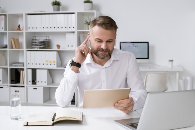 Successful employer with touchpad watching online video while sitting by desk in front of laptop