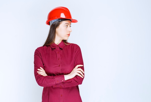 빨간색 하드 헬멧을 쓴 성공적인 여성 건축가가 서서 그녀의 옆을 바라보고 있습니다.