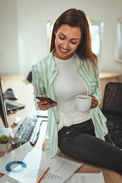 A successful female entrepreneur is writing messages on a smartphone and drinks coffee in the office.