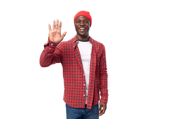 Успешный дружелюбный красивый черный американец, одетый в красную рубашку и кепку, машет рукой на белом