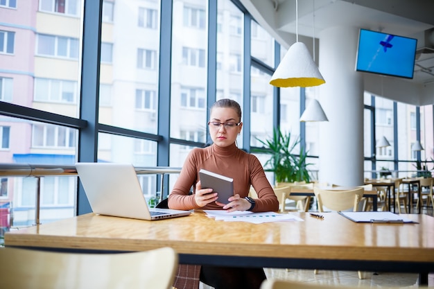 큰 창문이 있는 자신의 사무실에서 성공한 여자 매니저는 커피 한 잔과 함께 노트북으로 새로운 경제 개발 사업 계획을 세웁니다. 문서를 가진 비즈니스 우먼이 나무 테이블에 앉아 있다