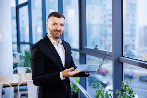 Successful handsome businessman sitting at his laptop next to\
large windows in modern office space