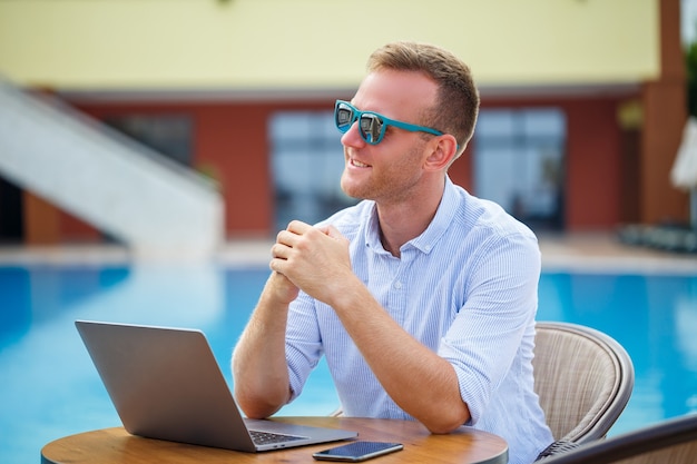 선글라스를 끼고 성공한 잘생긴 남성 사업가는 수영장 근처에 앉아 있는 노트북에서 일합니다. 원격 작업. 프리랜서