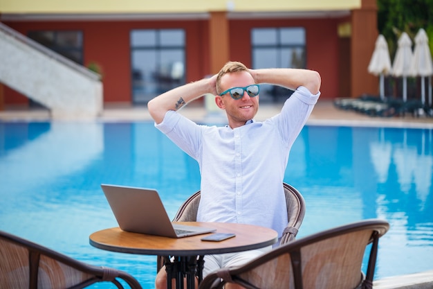 Successful handsome male businessman in sunglasses works at a laptop sitting near the pool. Remote work. Freelancer