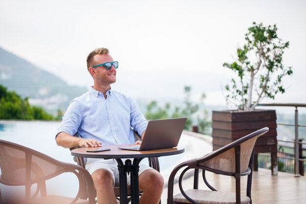 Successful handsome male businessman in sunglasses works at a laptop sitting near the pool. Remote work. Freelancer