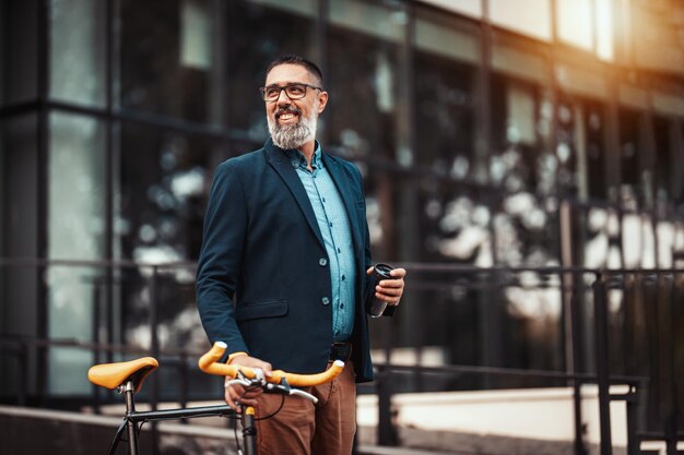 Successful handsome middle-aged businessman with a cup of coffee to go and with bicycle beside him, walks in front of office district.