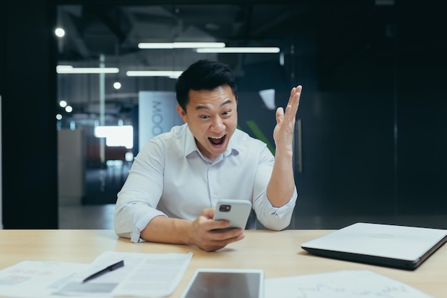 Uomo d'affari asiatico di successo e felice che celebra la vittoria seduto e lavorando in un ufficio moderno