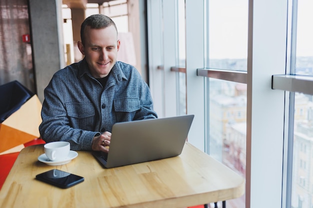 Successful happy business man is sitting at a table in a cafe, holding a cup of coffee and using a laptop.