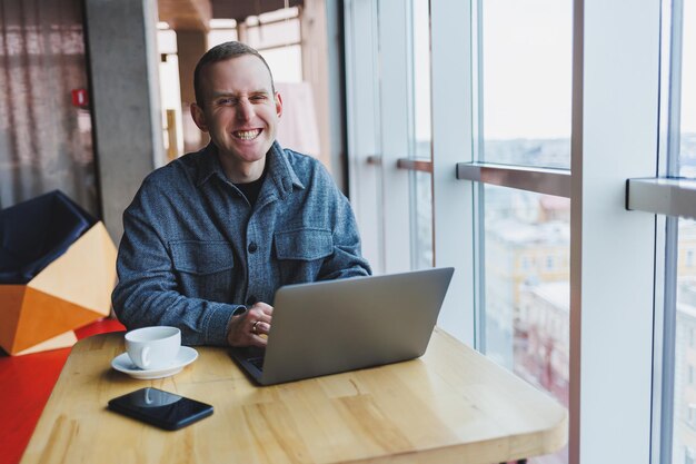 L'uomo d'affari felice di successo è seduto a un tavolo in un bar con in mano una tazza di caffè e utilizzando un computer portatile