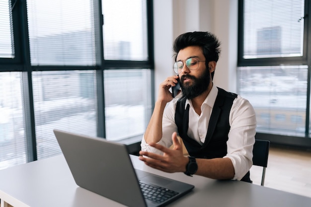 Successful Indian bearded business man in glasses working at laptop computer sitting at desk and talking on smartphone by window