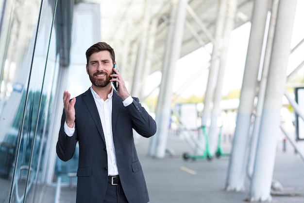 Successful male boss businessman talking outdoors near office uses cellphone confident smiles and rejoices
