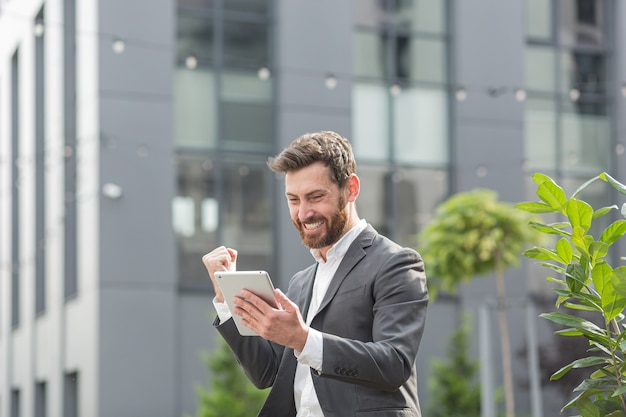 Successful male businessman got good win news reads them from a tablet computer, rejoices and thanks for the victory and success