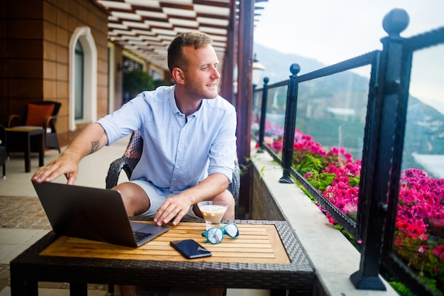 A successful male businessman is sitting at a table with a laptop, he is working on vacation. Vacation and remote work. The guy is sitting in a restaurant on a terrace with a panoramic view