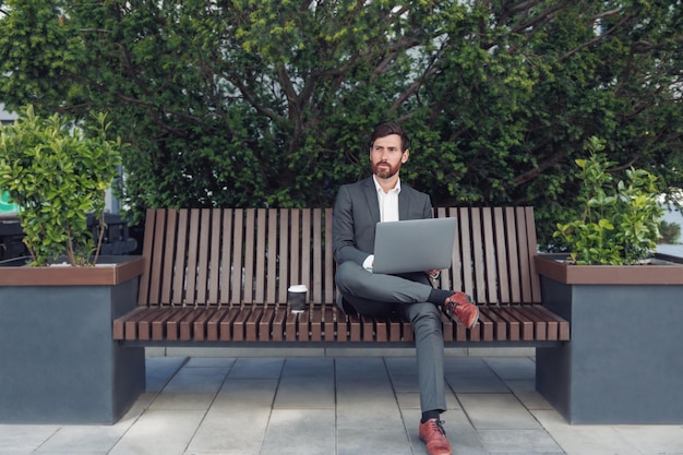 Successful male businessman working on laptop at lunchtime sitting on bench near office