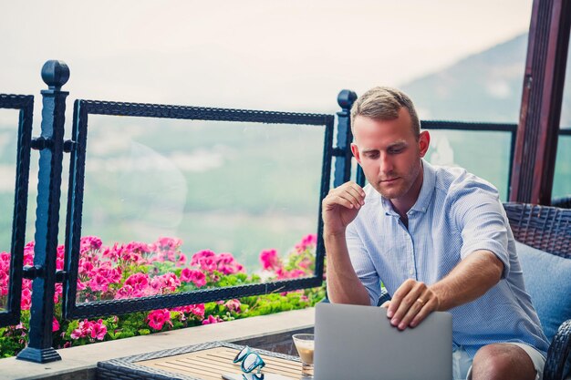 Successful male businessman working on vacation behind a laptop with a mountain view Online manager workflow Work outdoors with a beautiful view from the balcony