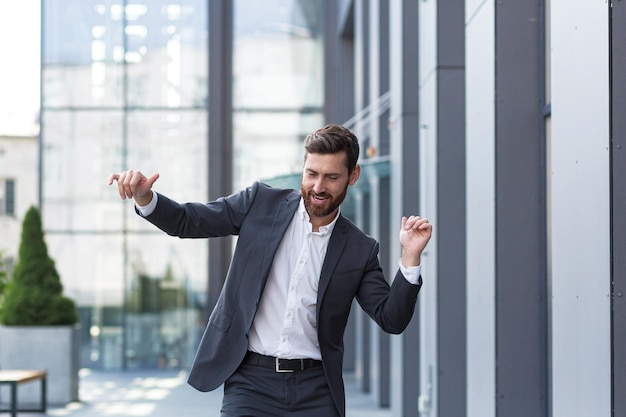 Успешный мужчина-фрилансер в деловом костюме танцует возле офиса, празднует выигрышную сделку и хорошее завершение рабочего дня