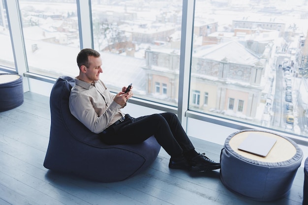 Successful male freelancer sitting in modern workspace and checking notifications on mobile phone in casual clothes Remote work