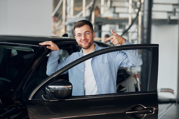 Successful man in glasses standing near brand new car indoors