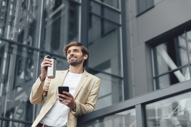 Uomo di successo in vestito elegante che tiene in una mano smartphone moderno e tazza di caffè in un altro