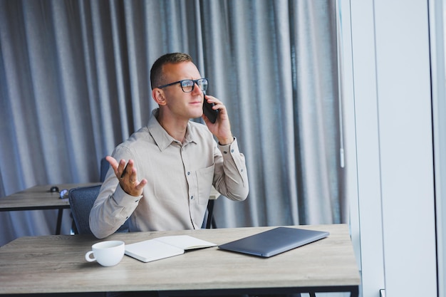 Successful man talking on smartphone and browsing netbook while surfing internet in office at table