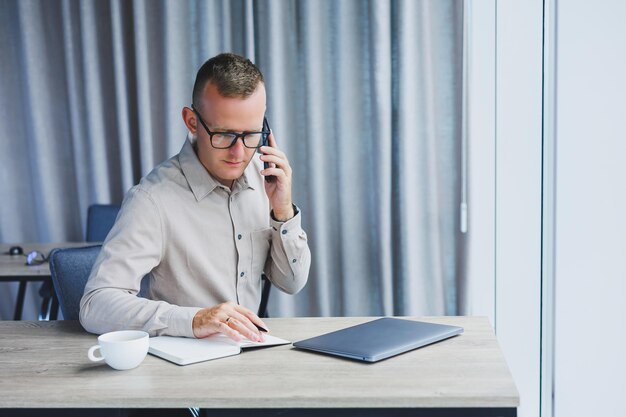 Успешный мужчина разговаривает по смартфону и просматривает нетбук во время серфинга в Интернете в офисе за столом