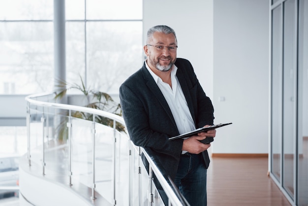 Successful manager. You can see it by his sincere smile. Photo of senior businessman in the spacious room with plants behind. Holding and reading documents