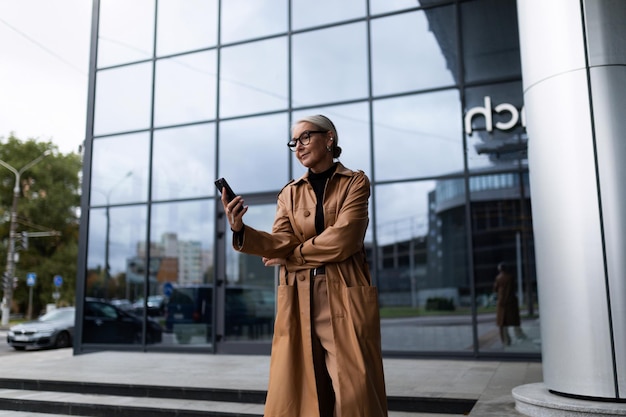 Successful mature adult caucasian woman looking at the phone at the entrance to the business center