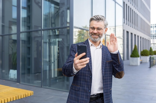 Successful mature man in business suit using app on phone for video call senior businessman outside