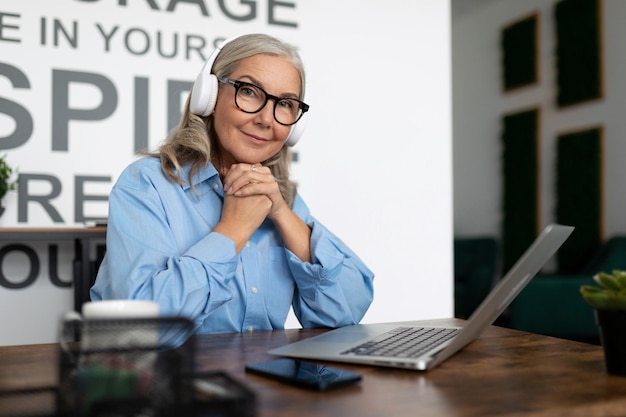 Successful mature woman manager watching training video on laptop with headphones sitting at table