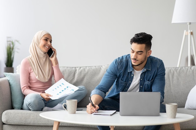 一緒に家からビジネスをしている成功した中東の家族、彼女の夫がラップトップで働いている間、スマートフォンで会話をしているドキュメントを持つヒジャーブの陽気な女性、コピースペース