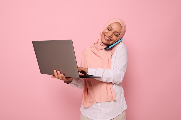 Successful Muslim Arab beautiful woman in hijab, dressed business smart casual talks on mobile phone , smiles looking at the monitor screen while typing text on a laptop. Copy space, pink background