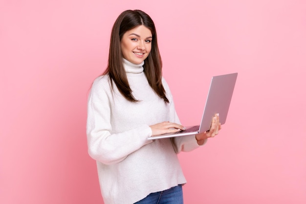 Successful office worker girl looking at camera with happy face holding laptop typing on keyboard wearing white casual style sweater Indoor studio shot isolated on pink background