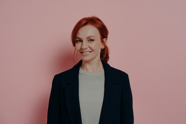 Successful pretty red-haired woman looking at camera with confidence and pleasant smile, standing isolated on rosy background in studio, stylish ginger female model in casual outfit smiling cheerfully