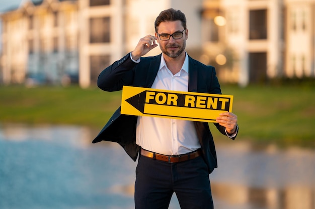 Successful real estate agent in a suit holding for rent sign near new apartment real estate agent wi