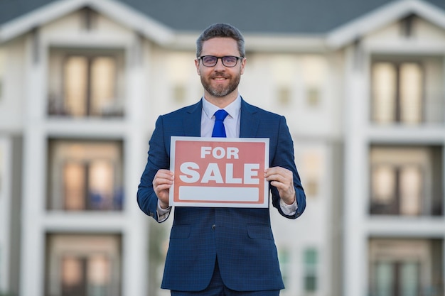 Successful real estate agent in a suit holding for sale sign near new apartment real estate agent wi