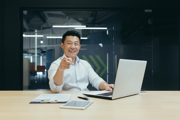 Успешный продавец азиатский бизнесмен счастлив и улыбается, глядя в камеру, указывая пальцем вперед человека