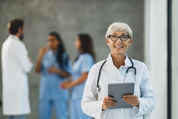 A successful senior female doctor looking at camera and holding digital tablet in a hospital hallway.