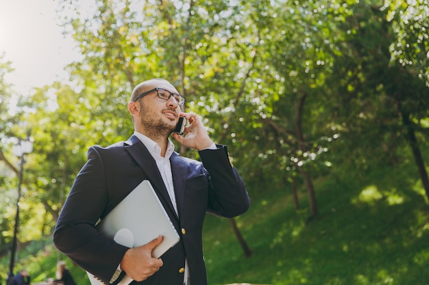 Successful smart businessman in white shirt, classic suit, glasses. Man stand with laptop pc computer, talk on mobile phone in city park outdoors on nature background. Mobile Office, business concept.