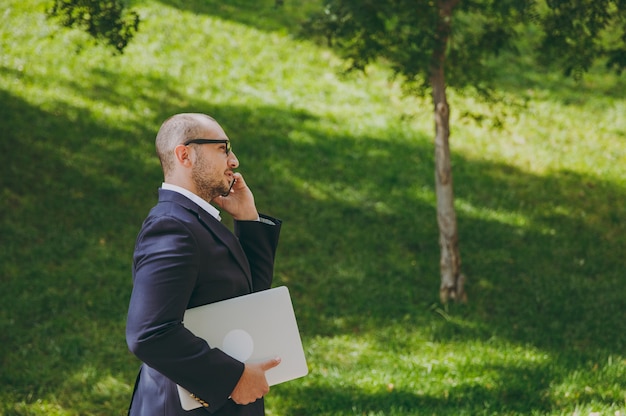 Successful smart businessman in white shirt, classic suit, glasses. Man stand with laptop pc computer, talk on mobile phone in city park outdoors on nature background. Side view, business concept.