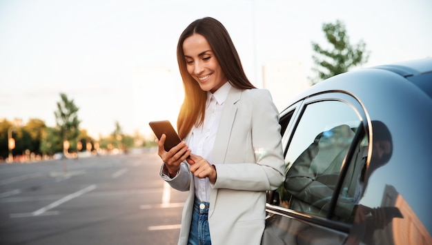 フォーマルなスマートウェアで成功した笑顔の魅力的な女性は、屋外の現代の車の近くに立っている間、彼女のスマートフォンを使用しています