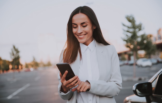 Successful smiling attractive woman in formal smart wear is using her smart phone while standing near modern car outdoors