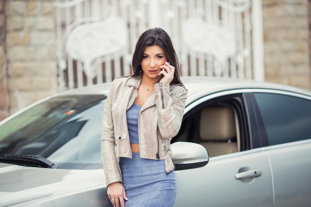 フォーマルなスマートウェアで成功した笑顔の魅力的な女性は、屋外の現代の車の近くに立っている間、彼女のスマートフォンを使用しています