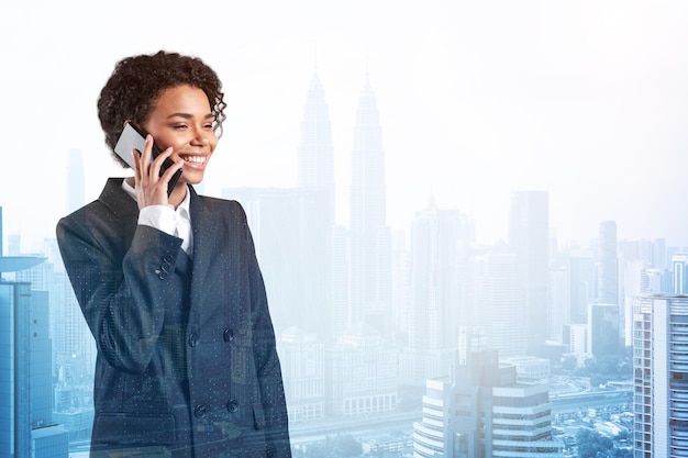 Успешная улыбающаяся чернокожая афроамериканская деловая женщина в костюме задумчиво разговаривает по телефону Городской пейзаж Куала-Лумпура Концепция консультантов как решателей проблем Небоскребы KL Двойная экспозиция