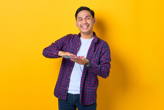 Успешный улыбающийся молодой азиат в клетчатой рубашке с банкнотами на желтом фоне