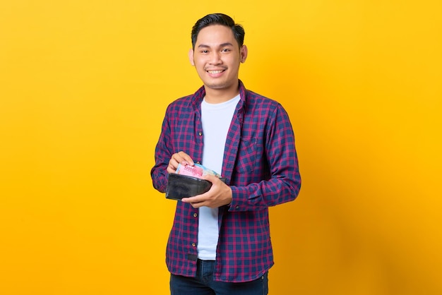 Successful smiling young Asian man in plaid shirt showing wallet full of money banknotes isolated on yellow background