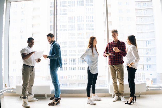 Successful team of young multiethnic startup business people discuss project together reading documents and using digital tablet standing on background of window and skyscrapers