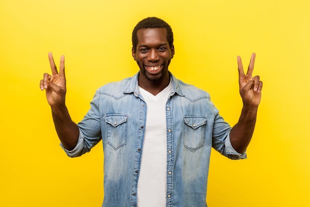 Successful winner. Portrait of excited man with toothy smile in denim casual shirt standing with raised hands and showing v sign or peace, victory gesture. studio shot isolated on yellow background