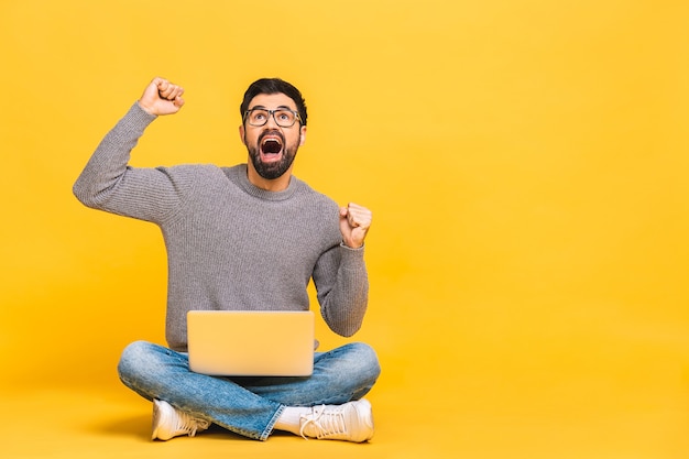 Successful winner! Portrait of a happy young bearded man using laptop and celebrating success or victory isolated over yellow background.