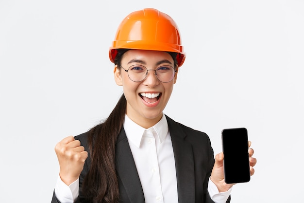 Successful winning asian female engineer, architect in safety helmet and suit, showing smartphone screen, fist pump and shouting yes from rejoice, triumphing over achievement, white background